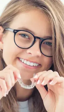 Smiling girl wearing glasses holding clear aligner tray for orthodontic treatment.
